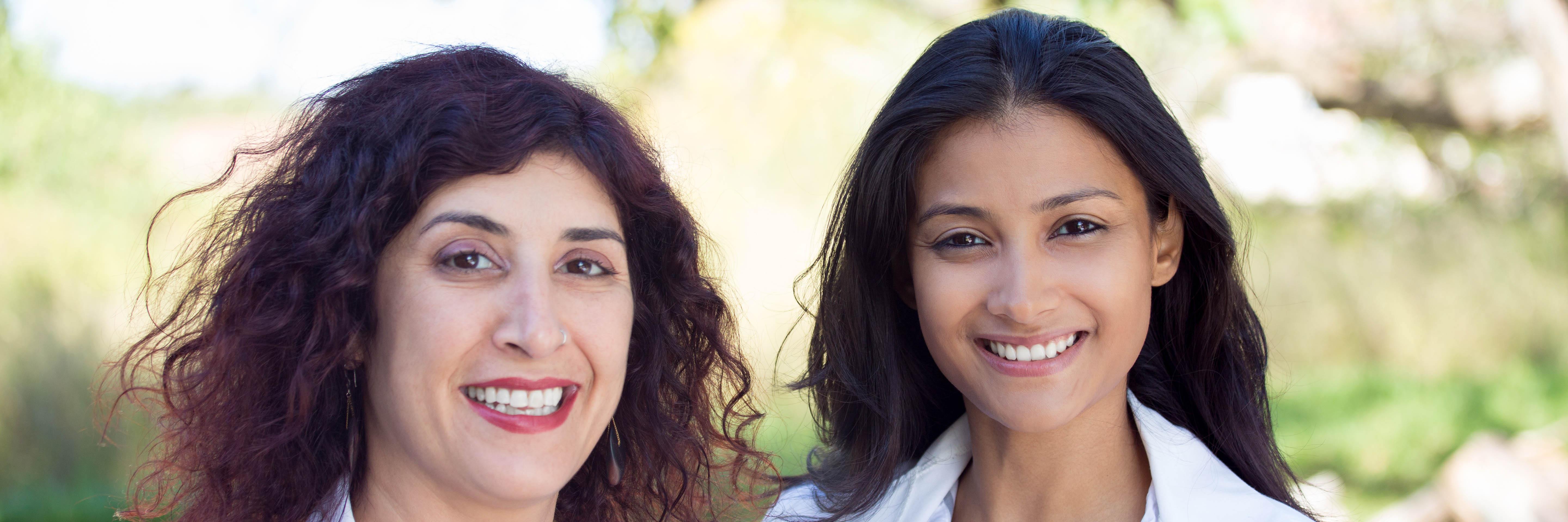 Two women clinicians, smiling, in white lab coats outdoors on a sunny day with trees in the background.